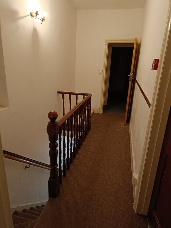 Hallway with wooden staircase railing, cream walls, brown carpet, and wall sconce lighting