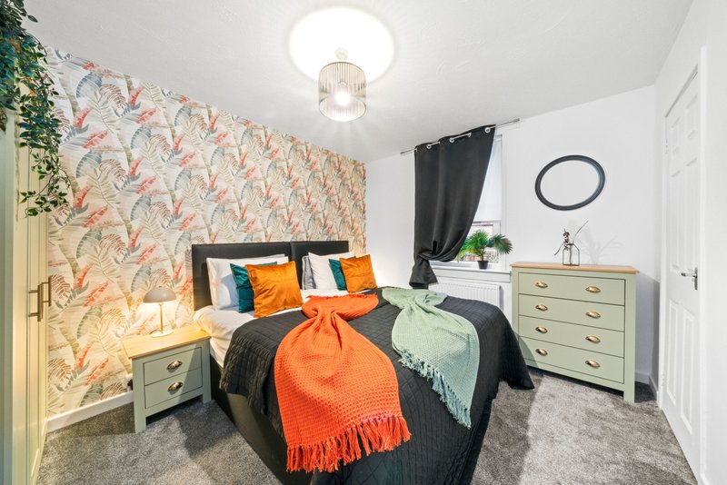 Bedroom with patterned wallpaper, double bed with orange throw, wooden dresser and bedside table
