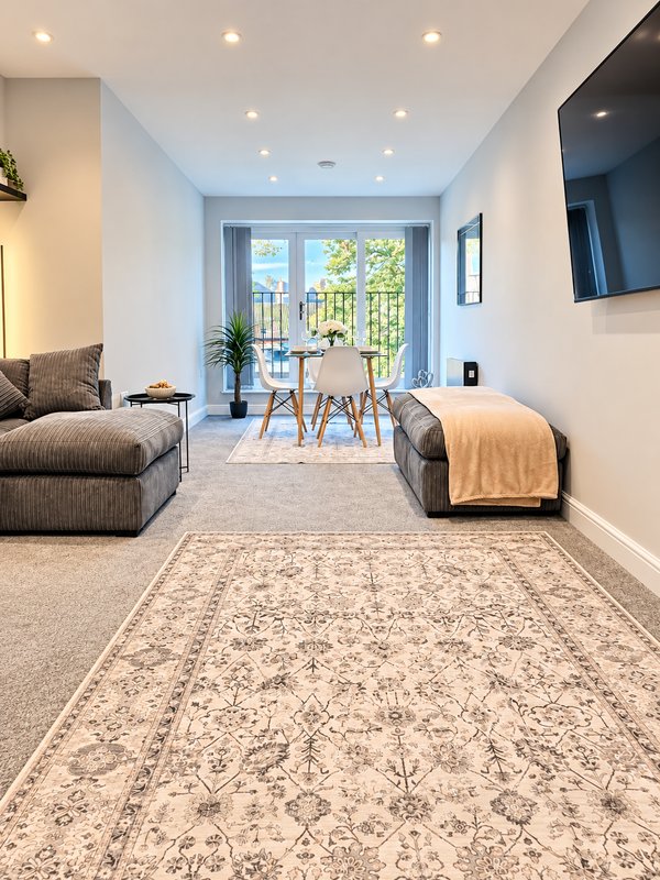 Modern living room with seating area, dining table, large windows overlooking balcony, mounted TV and recessed lighting