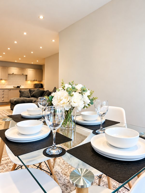Modern dining table set with white plates, glasses and fresh flowers, kitchen visible behind