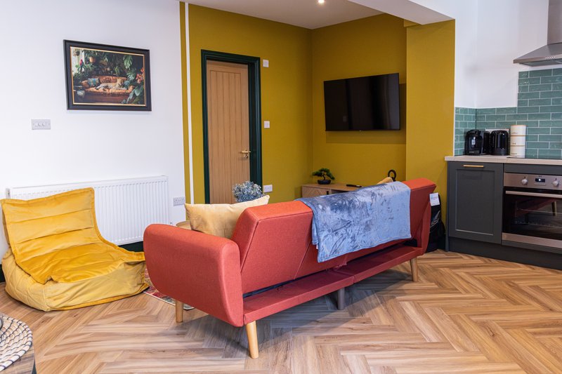 Open-plan living area with red sofa, yellow bean bag, kitchen, and mounted TV on mustard wall