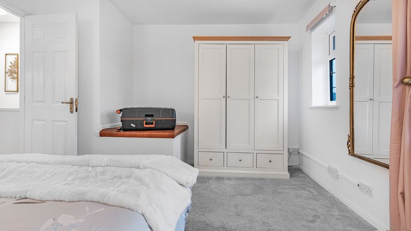 Bright bedroom with double bed, white wardrobe, luggage shelf and grey carpet flooring