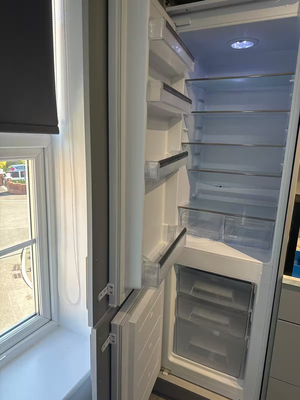 Empty open refrigerator with shelves and freezer drawer, window visible on left