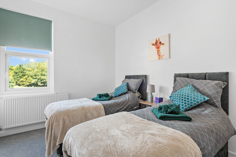 Twin bedroom with grey upholstered headboards, white and beige bedding, turquoise pillows, and window with green blind. Can be made in to a king bed.