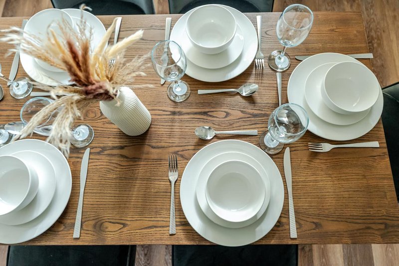 Wooden dining table set for four with white plates, glassware, and dried pampas grass centerpiece