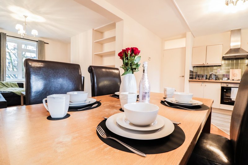 Modern dining area with wooden table, place settings, red flowers, and kitchen visible in background