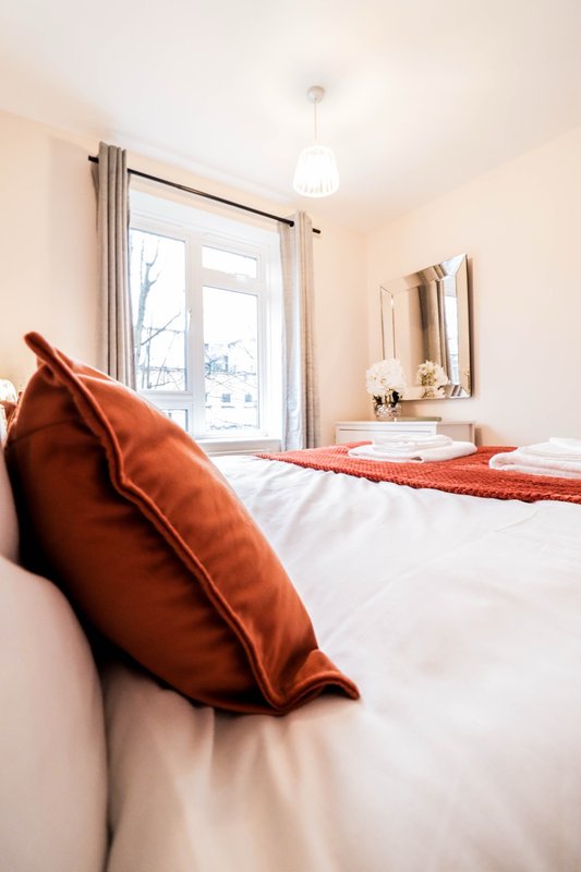 Bright bedroom with double bed, orange cushions, window with curtains and mirror on wall