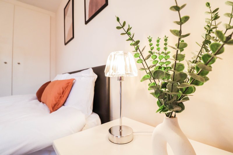Bedroom with white bed, orange cushion, bedside lamp, and potted plants on nightstand