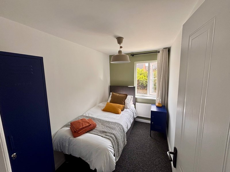 Compact bedroom with single bed, yellow cushions, dark blue door and window with garden view