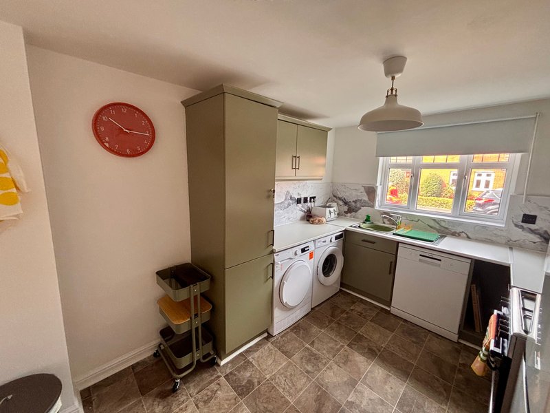 Kitchen with white appliances, beige cabinetry, red wall clock, and window with autumn view