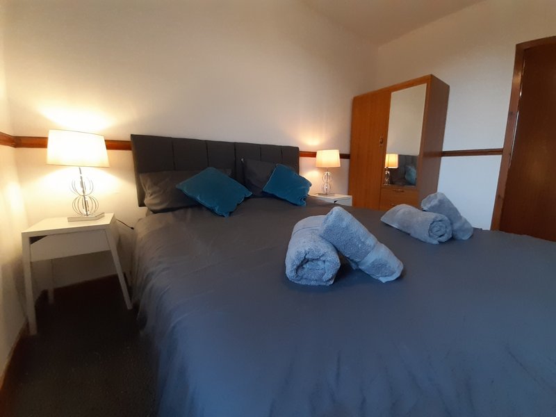 Bedroom with double bed, blue pillows, rolled towels, bedside lamps and wooden wardrobe
