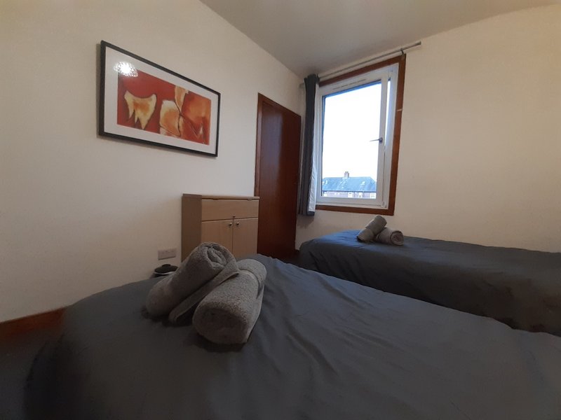 Small bedroom with two single beds, wooden dresser, framed artwork and window with city view