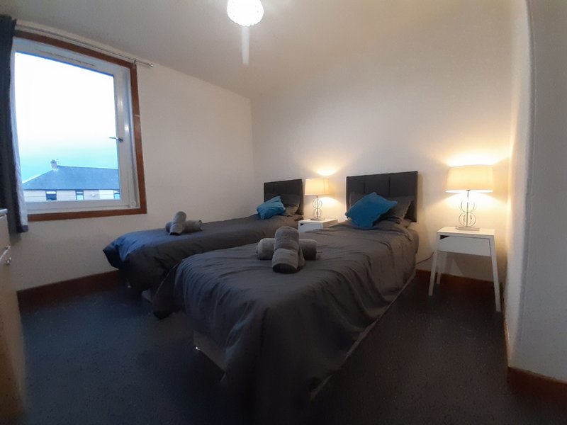 Bedroom with two single beds, dark bedding, blue cushions, bedside lamps and window view
