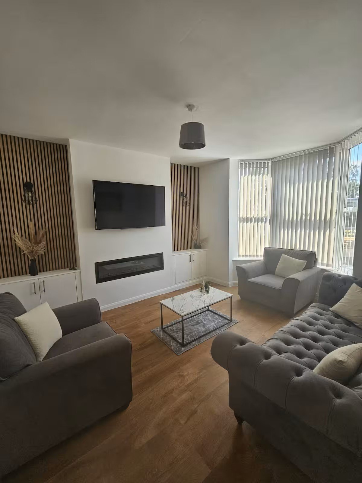Modern living room with gray upholstered seating, wall-mounted TV, fireplace, wooden flooring and large windows