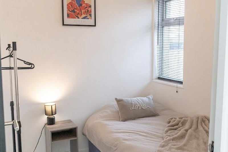 Small bedroom with single bed, bedside lamp, window with blinds and framed artwork