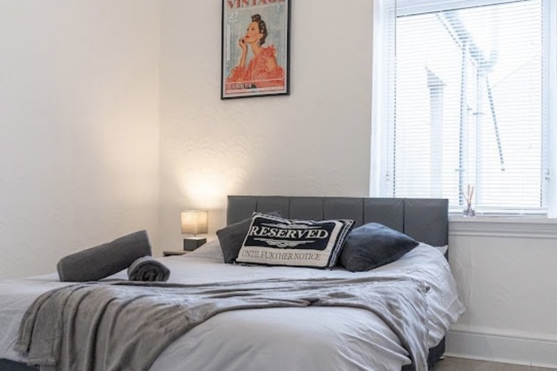 Modern bedroom with gray upholstered headboard, unmade bed with pillows and throws, bedside lamp and window.