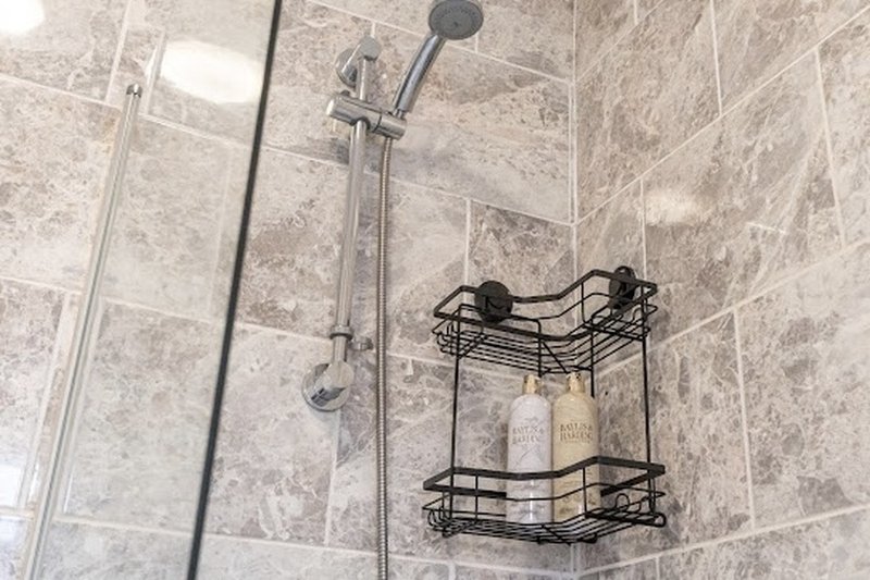 Bathroom shower enclosure with grey tile walls, chrome showerhead and black wire shelving holding toiletries