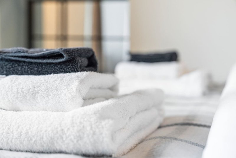Folded white and dark towels stacked on bed with windows in background