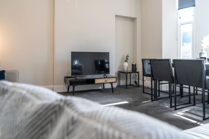 Modern living room with TV on wooden stand, dining chairs, and bright natural light from windows