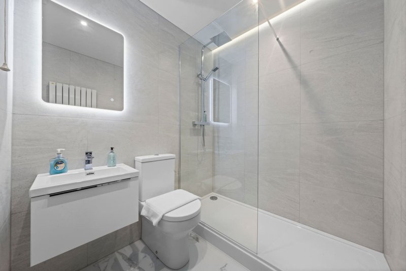 Modern bathroom with illuminated mirror, white vanity, toilet, and walk-in shower with glass partition