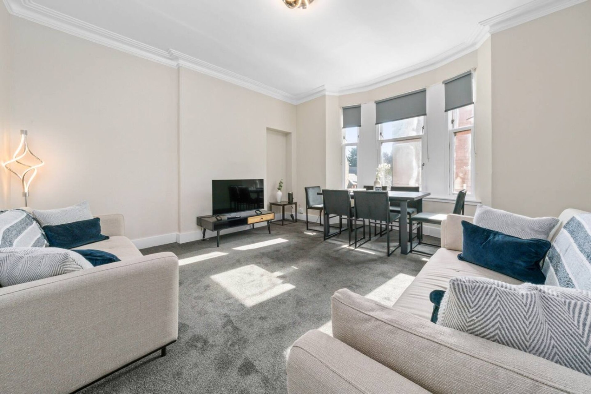 Bright living room with cream sofas, dining table, TV, large windows and geometric cushions