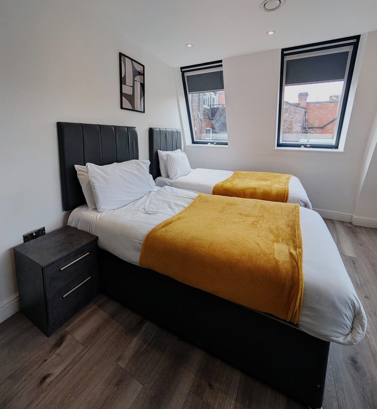 Modern bedroom with two single beds, black headboards, gold throws, white pillows, and windows with city views