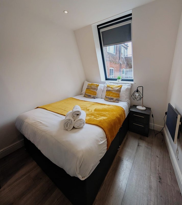 Small bedroom with double bed, yellow bedding, white pillows, black nightstand and window