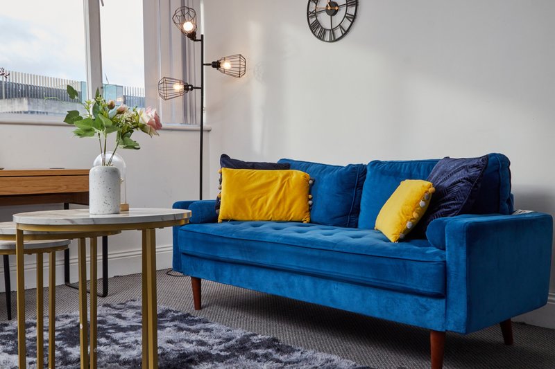 Modern living room with blue velvet sofa, yellow cushions, wooden side table with flowers and floor lamp