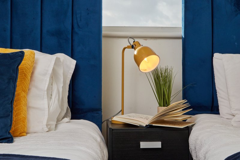 Twin bedroom with blue upholstered headboard, white bedding, gold lamp and open book on nightstand