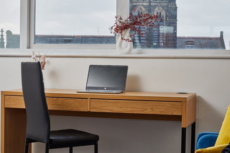 Home office desk with laptop, wooden drawers, black chair, and city skyline view through window