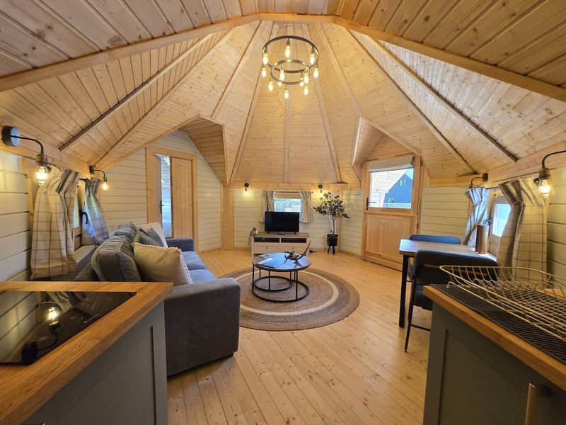 Open-plan glamping interior with wooden ceiling, sofa, kitchenette, dining table and pendant lighting