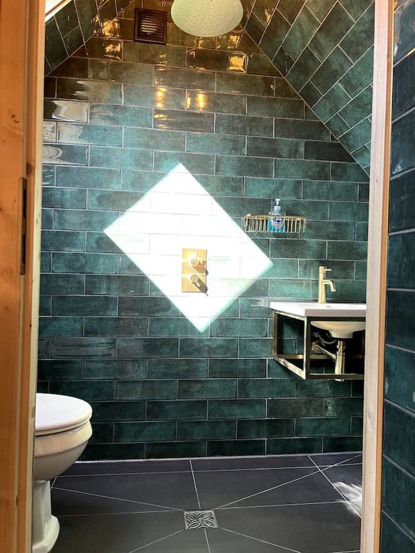 Bathroom with dark green subway tile walls, white diamond-shaped mirror, toilet, and pedestal sink