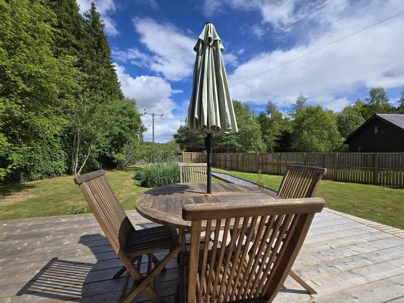 Garden patio with wooden dining table, chairs, and closed parasol under blue sky