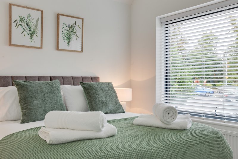 Bright bedroom with grey upholstered headboard, green and white bedding, rolled towels, and botanical wall art