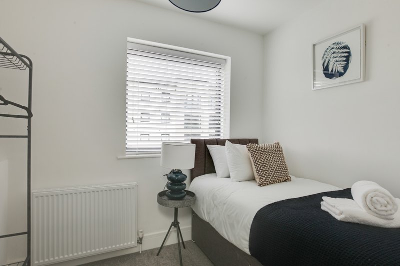 Modern bedroom with double bed, white pillows, dark bedding, small side table with decorative object, and window with blinds