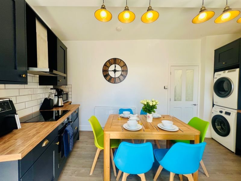 Modern kitchen-diner with dark cabinetry, wooden table, bright chairs and stacked washing machines