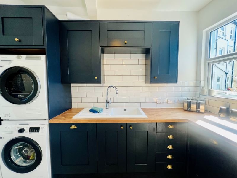 Modern kitchen with dark blue cabinetry, white subway tile backsplash, wooden countertop, and stacked washing machines