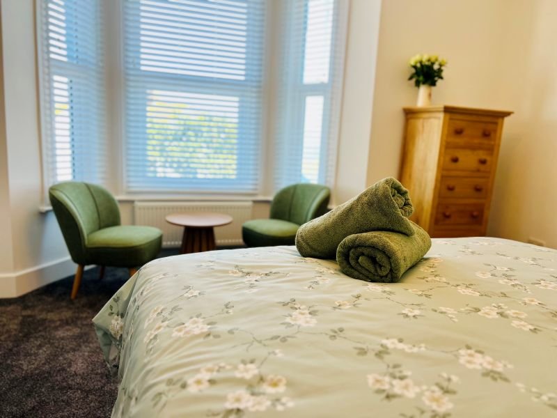 Bedroom with double bed, green armchairs, wooden dresser and bay windows with shutters