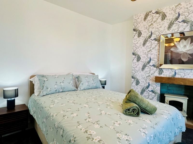 Bedroom with double bed, patterned bedding, wooden nightstand, lamp, and fireplace with mirror