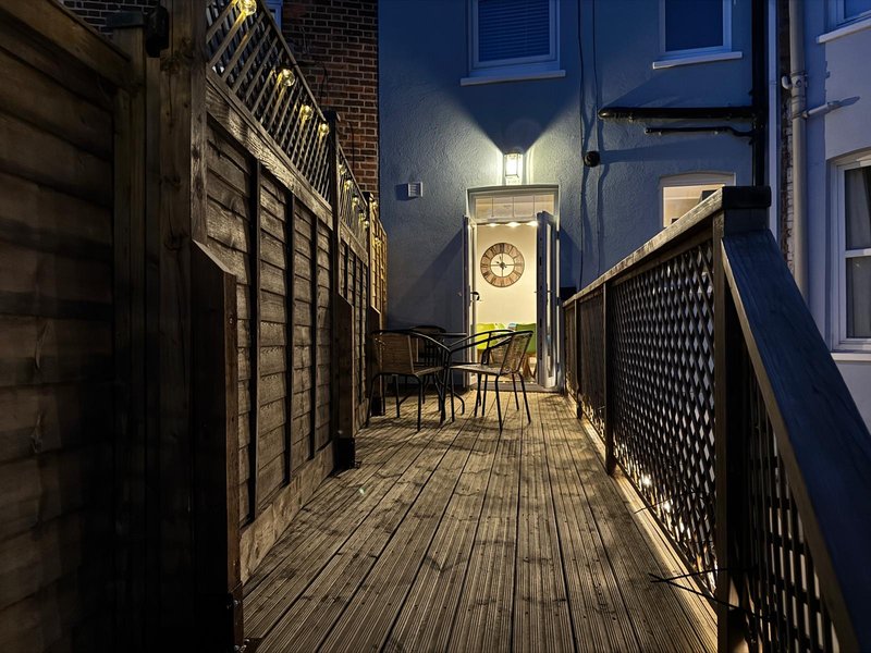 Narrow exterior alleyway with wooden deck, brick walls, chairs, and illuminated doorway at night