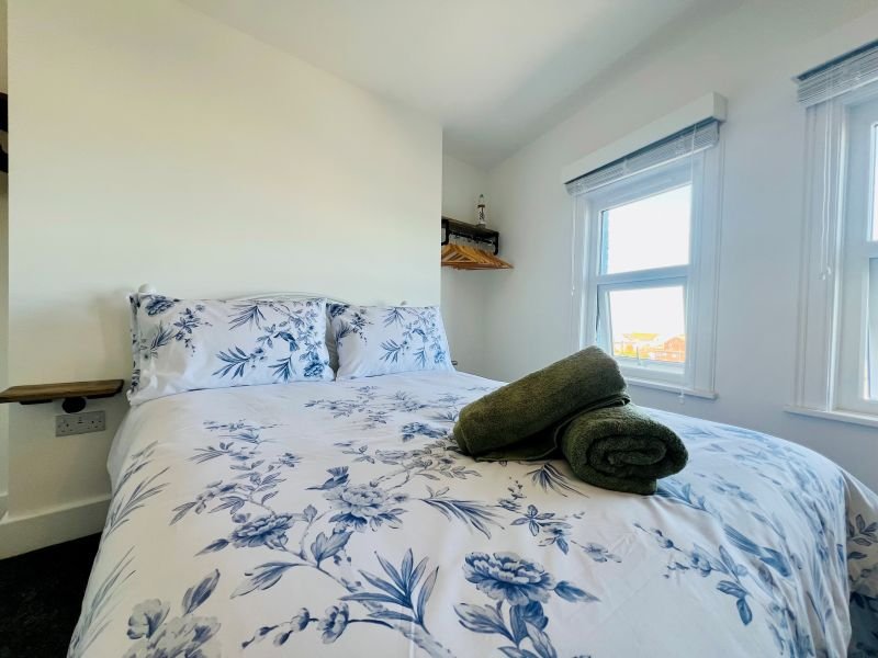 Bright bedroom with double bed, blue floral bedding, green towels, and windows with light curtains