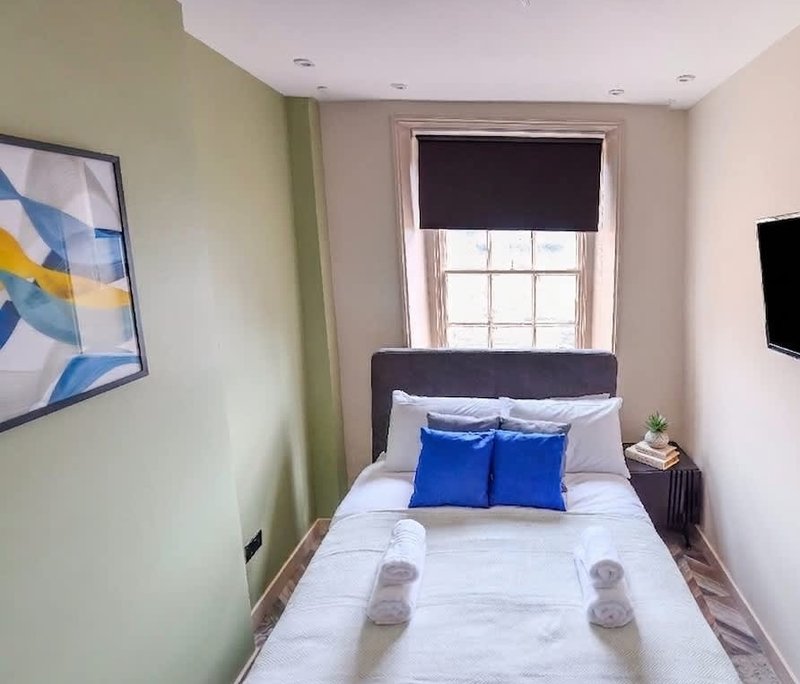 Compact bedroom with single bed, blue pillows, window with blind and framed artwork