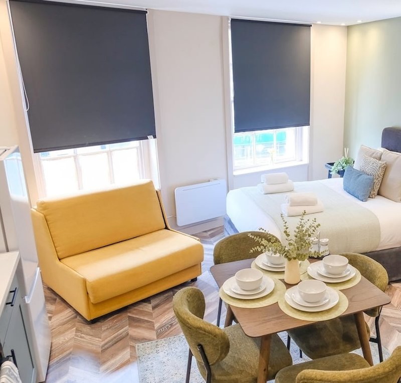 Studio apartment with yellow chair, dining table, bed, and blue roller blinds