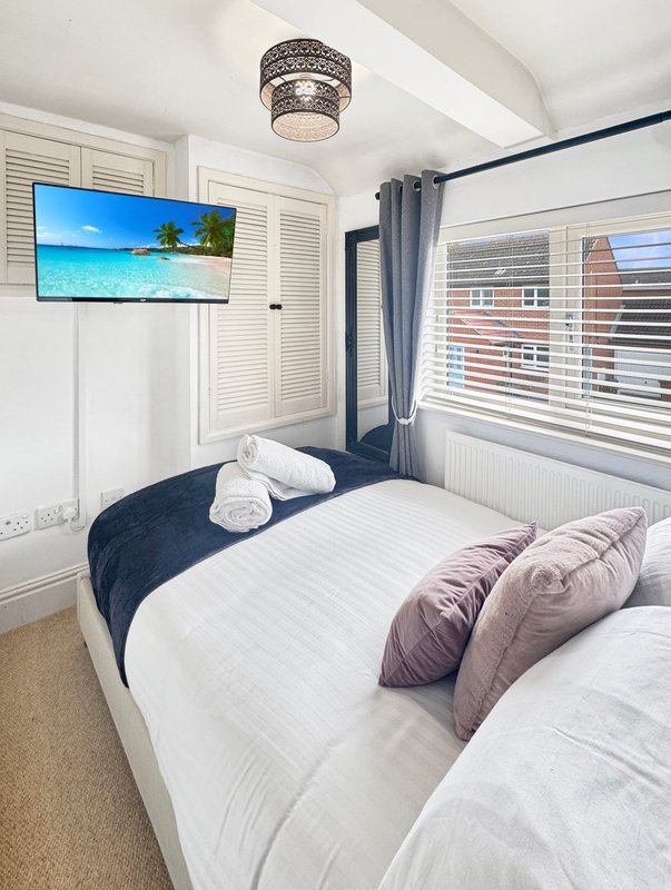 Bedroom with double bed, rolled towels, purple pillows, wall-mounted TV, ceiling light and window blinds