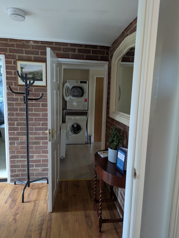 Hallway with brick wall, wooden flooring, washing machines visible through open door, and side table with decor