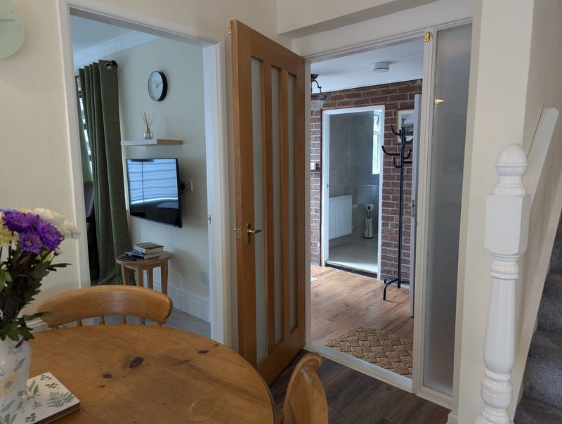 Open-plan living area with wooden dining table, purple flowers, TV mounted on wall, and doorway to hallway