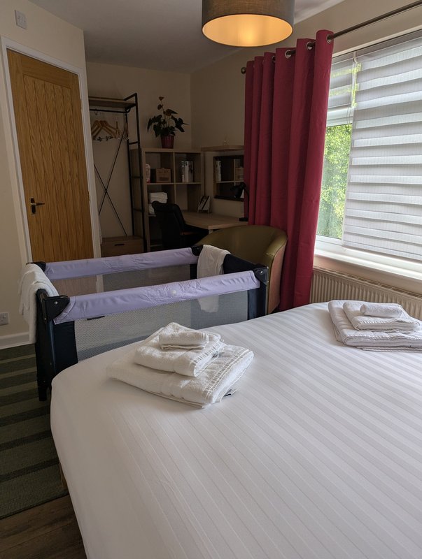Bedroom with double bed, travel cot, desk, burgundy curtains and window blinds