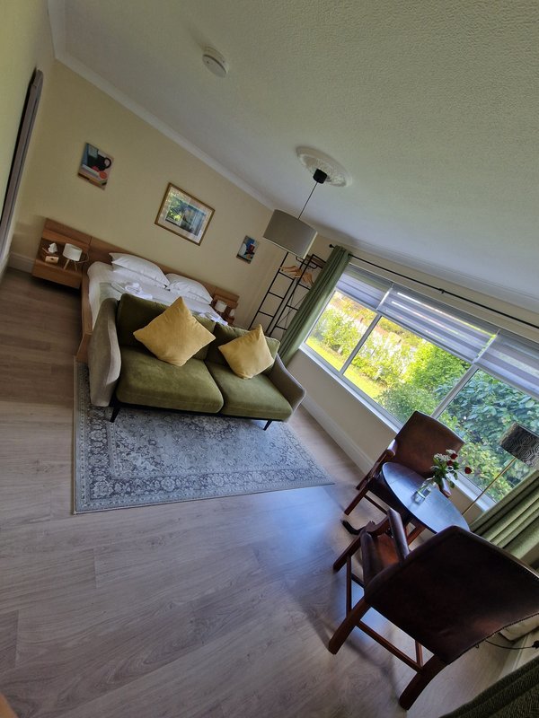 Bright living room with olive sofa, wooden chairs, large window overlooking garden, and framed wall art
