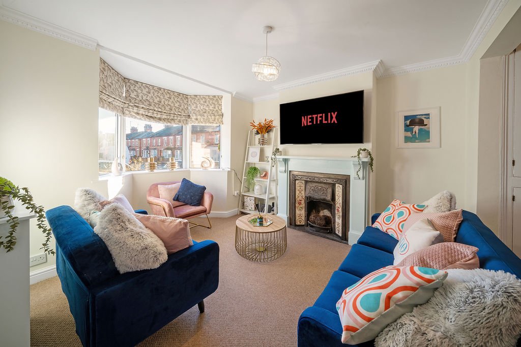 Bright living room with two blue sofas, fireplace, wall-mounted TV, and large bay window