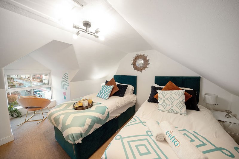 Bright attic bedroom with two single beds, teal bedding, cushions, and a seating area by the window